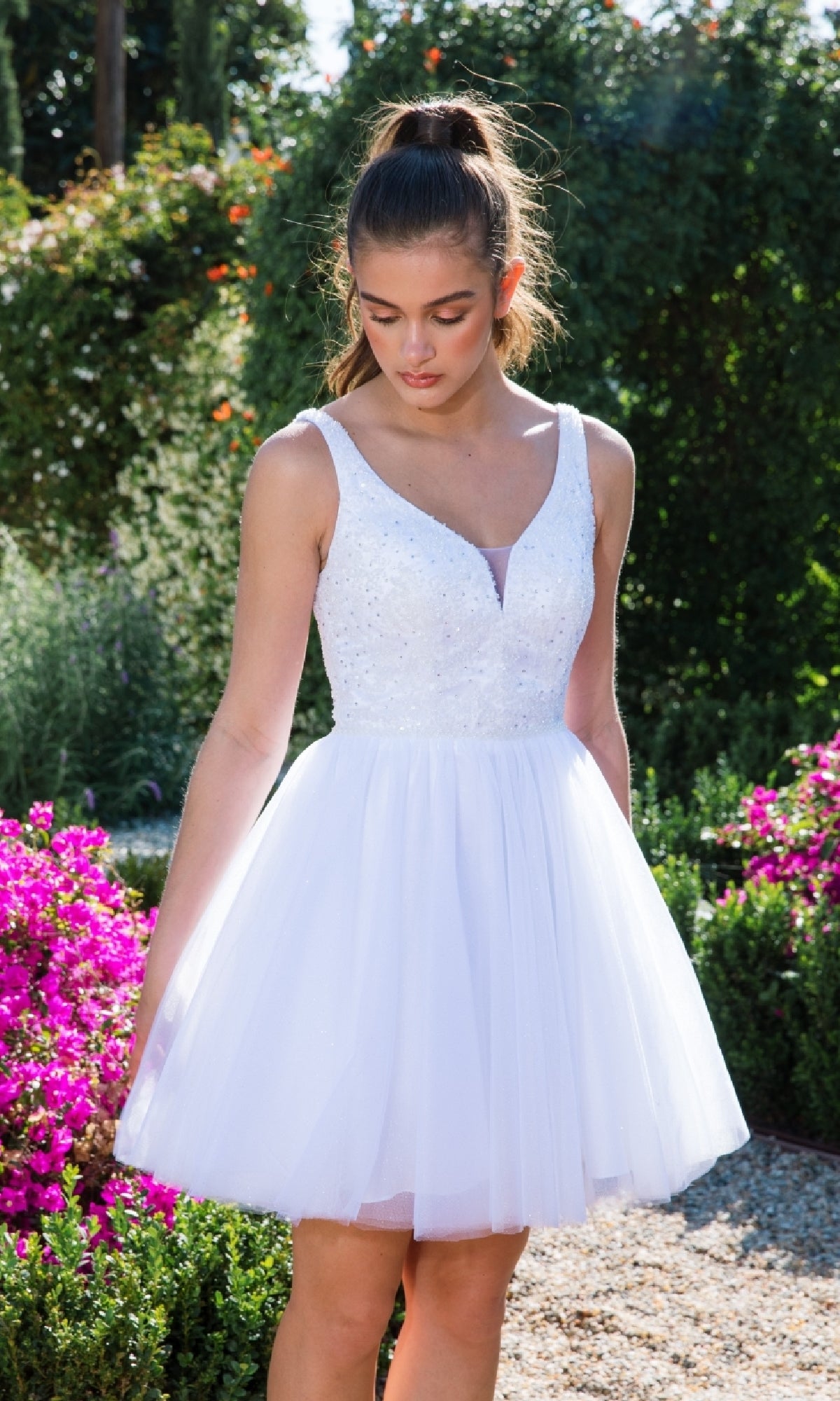 A young woman wears the Glitter-Bodice Short A-Line Hoco Dress: Eureka 9926, featuring a sleeveless white bodice and tulle skirt, as she stands in a blooming garden surrounded by lush greenery.